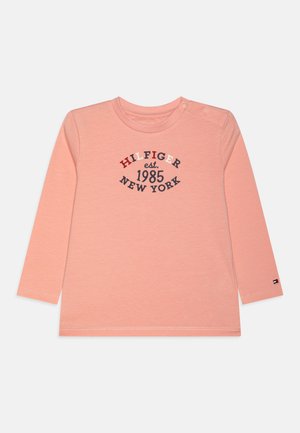 Ilgais rankovėmis, šviesiai rožinis marškinėliai su apvaliu kaklu, puikuojasi jūriniais ir raudonais užrašais: "HILFIGER est. 1985 NEW YORK," ir mažas logotipas ant rankovės.