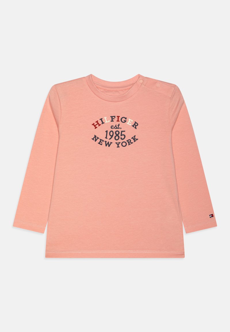 Långärmad, ljusrosa t-shirt med rund halsringning, pryds av marinblå och röda tryckta texter: "HILFIGER est. 1985 NEW YORK," och en liten logotyp på ärmen.