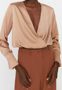 Indpakket bluse i blød beige satin med lange ærmer, dyb V-hals og løs pasform, parret med skræddersyede brune bukser med knaplukning.