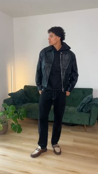 Νεαρός άνδρας που φοράει μαύρο δερμάτινο μπουφάν, μαύρη κουκούλα, μαύρο παντελόνι και καφέ μοκασίνια, στέκεται σε σαλόνι με πράσινο καναπέ και φυτά.
