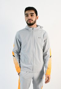 Chaqueta deportiva de color gris claro con acentos naranjas, rayas negras y capucha. Cuenta con un cierre frontal completo y un diseño texturizado en las mangas.