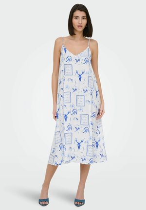 Femme debout dans une robe midi blanche avec des imprimés fruits de mer bleus et encadrements, bretelles spaghetti et talons ouverts bleus assortis sur un fond clair.