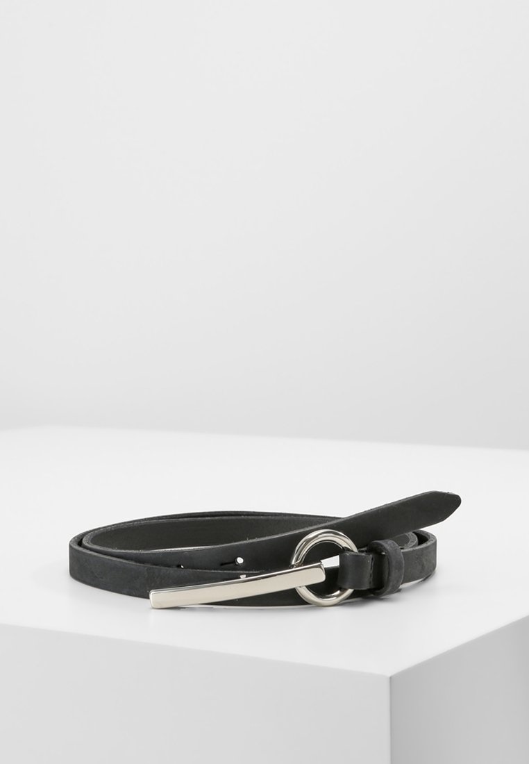Ceinture en cuir noir avec une boucle circulaire en métal argenté et une épingle métallique droite, enroulée sur une surface blanche.