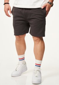 Dunkelgraue Sportshorts aus einem Baumwollmix, ausgestattet mit einem elastischen Taillenbund und Seitentaschen. Kombiniert mit weißen Turnschuhen und gestreiften Socken.