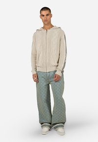 Πλέκτο μπεζ φούτερ με φερμουάρ και υφή διαμαντιού, με γκρι διακοσμητικές ρίγες. Φορεμένο με ανοιχτό μπλε τζιν με σχέδιο και φαρδιά γραμμή και λευκά παπούτσια.