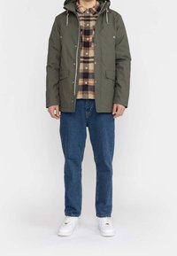 Olivgrön huva jacka med tryckknappar, två framfickor, tillsammans med en rutmönstrad flanellskjorta, blå denimjeans och vita sneakers.