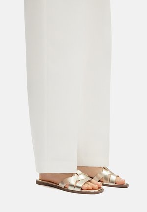 Metaliczne złote sandały z krzyżującymi się paskami, płaską podeszwą i ozdobnym wzorem. Noszone z luźnymi, białymi spodniami.