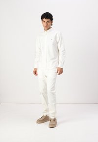 Hvit langermet skjorte med knapper og vertikale striper, kombinert med hvite bukser og beige sko. Enkel design med krage.