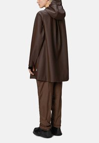 Giacca impermeabile marrone con cappuccio, design lungo e superficie liscia, abbinata a pantaloni marroni coordinati e scarpe nere.
