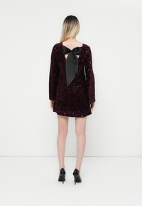 Rochie scurtă bordeaux cu mâneci lungi, acoperită cu paiete, având un decupaj adânc la spate legat cu o fundă din satin negru. Tocuri negre.