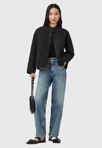 Giubbotto nero trapuntato con dettagli a intaglio, abbinato a jeans a vita alta blu chiaro e scarpe nere basse. Modella che tiene una borsa nera.