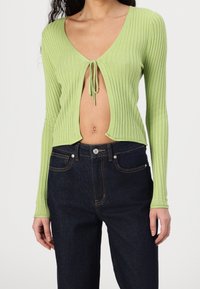 Grünes geripptes Strickoberteil mit tiefem V-Ausschnitt und einer Bindeschleife in der Mitte, kombiniert mit hochgeschnittenen dunklen Jeans.
