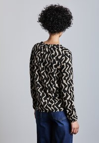 Maglietta a maniche lunghe con un motivo zigzag nero e beige, scollo rotondo, texture morbida e dettaglio arricciato sul retro. Indossata con jeans blu.