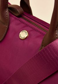 Lähivõte lillast kangast kotist, millel on pruunid tekstuuriga sangad, metallist lukk ja ümar kuldne nööp, millele on graveeritud "Joules" logo.