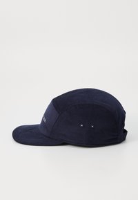 Tumedate nööbist müts ümarate kroonidega, lame ääre ja kahe ventilatsiooniavaga külgedel. Esiküljel on tikandiga logo.