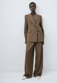 Bruine dubbelgeknoopte blazer met twee zakken, gecombineerd met wijde broekspijpen. Glad textuur, minimalistisch design en lichtgekleurde schoenen.