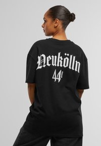 Čierne oversized tričko s bielym gotickým písmom "Neukölln" a vytlačeným číslom "44" pod ním. Má voľný strih a krátke rukávy.