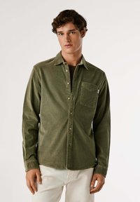 Camisa de pana verde oliva con cuello, cierre frontal con botones, un bolsillo en el pecho y botones en un acabado metálico claro.