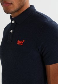 Námořnická modrá polo triko s texturovanou tkaninou, tři knoflíky na zapínání a červené vyšívané logo na hrudi. Krátké rukávy a klasický límec.