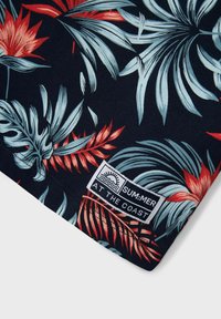 Camisa de tejido de manga corta con un fondo azul marino oscuro y vibrantes patrones florales tropicales en rojo y azul claro. La etiqueta dice "Verano en la Costa".
