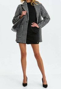 Svartvit hundtandsmönstrad kappa med långa ärmar och krage, kombinerad med en figurnära svart topp och kjol, svart handväska och svarta klackar.