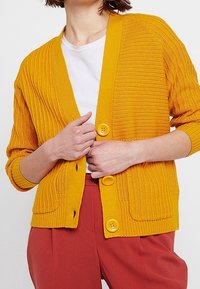 Žlutý žebrovaný cardigan se třemi knoflíky, otevřeným předním dílem a dlouhými rukávy, nošený přes bílou košili. Texturovaná pletená látka.