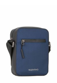 Sac bandoulière en tissu bleu de forme rectangulaire, accents noirs, fermeture éclair argentée et logo embossé à l'avant. Dispose d'une bandoulière réglable.