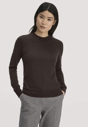 Jonge vrouw met lang zwart haar die een donkerbruine trui en grijze broek met patroon draagt, staand met haar handen in haar zakken tegen een effen achtergrond.