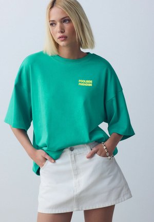 Mladá žena so svetlými bob vlasmi, oblečená vo voľnej zelenej košeli s nápisom "Poolside Paradise" a bielej rifľovej mini sukni, ruky v vreckách.