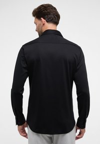 Schwarz langärmliges Hemd, glattes Material, Standardkragen, Rückenpassform, geknöpfte Manschetten, abgerundeter Saum, getragen mit hellgrauten Hosen.