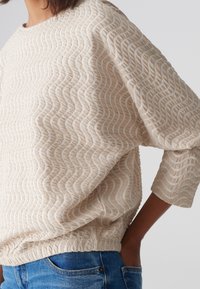 Haut beige texturé avec un motif ondulé, manches trois quarts et une taille cintrée, associé à un jean bleu.