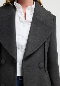 Abrigo de lana gris con un diseño de doble botonadura, grandes botones redondos y un cuello ancho, superpuesto a una camisa blanca de botones.