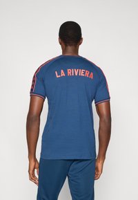 Granatowy t-shirt z krótkim rękawem, z napisem "LA RIVIERA" w czerwonym kolorze na plecach, z kontrastowymi czerwonymi paskami na ramionach.
