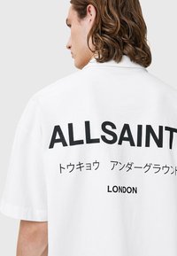 Hvit bomulls t-skjorte med en stor svart "ALLSAINTS" logo på baksiden, med tilleggstekst i ulike skrifttyper og "LONDON" nederst.