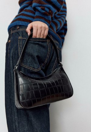 Sac à main en cuir embossé façon croco noir, de forme incurvée avec une poignée courte, tenu par une main près de la poche arrière d'un jean en denim bleu.