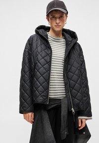 Quiltota melna pūkainā jaka ar kapuci, kura ir aprīkota ar rāvējslēdzēja aizdari priekšpusē, kopā ar pelēku svītrainu garām piedurknēm krekla un tumšiem biksēm.