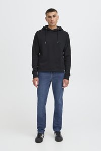 Ung man stående klädd i svart hoodie, blå jeans och svarta sneakers med vita ränder mot en enfärgad vit bakgrund.