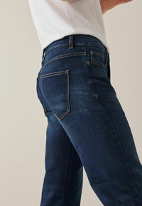 Sötétkék farmernadrág, slim fit szabással, öt zsebes kialakítással és kontrasztos narancssárga varrással. Simás textúra, enyhén kopott hatással a combokon.