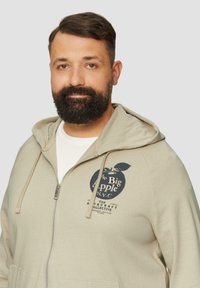 Beige rits hoodie met voorzakken en verstelbare capuchon. Heeft een donkerblauwe grafische print van een appel en de tekst "The Big Apple NYC."