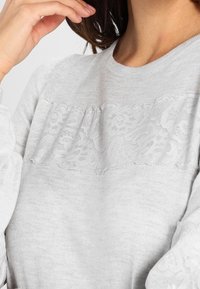 Felpa grigia chiara con un pannello in pizzo testurizzato sul petto, scollatura rotonda e dettagli arricciati sotto il pizzo.