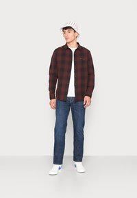 Camisa de franela a cuadros rojos y negros, camiseta blanca de cuello redondo, jeans azules, zapatillas blancas con detalles en azul y un sombrero de cubo a rayas.