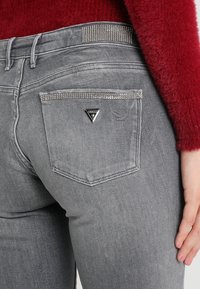 Gros plan d'une personne portant un jean gris avec des détails en strass sur la poche arrière et la ceinture, accompagné d'un pull rouge pelucheux.