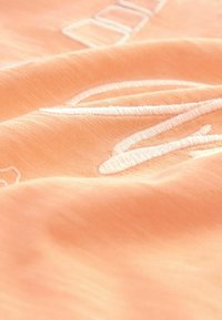 Gros plan sur un tissu couleur pêche avec des lettres blanches brodées en relief et des plis doux.