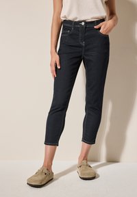 Jeans de tobillo de mezclilla azul oscuro con costuras de contraste claras, diseño de cinco bolsillos y forma ajustada. Combinados con zapatos beiges de slip-on.