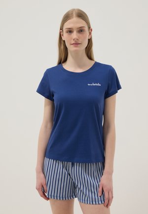 Mujer joven vistiendo una camiseta azul de manga corta con texto blanco pequeño y pantalones cortos a rayas verticales azules y blancas, de pie sobre un fondo neutro.