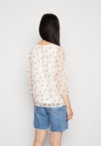 Blusa bianca con delicata stampa floreale, tessuto trasparente, maniche a tre quarti, scollatura rotonda e un piccolo bottone sul retro. Abbinata a pantaloni shorts in denim.