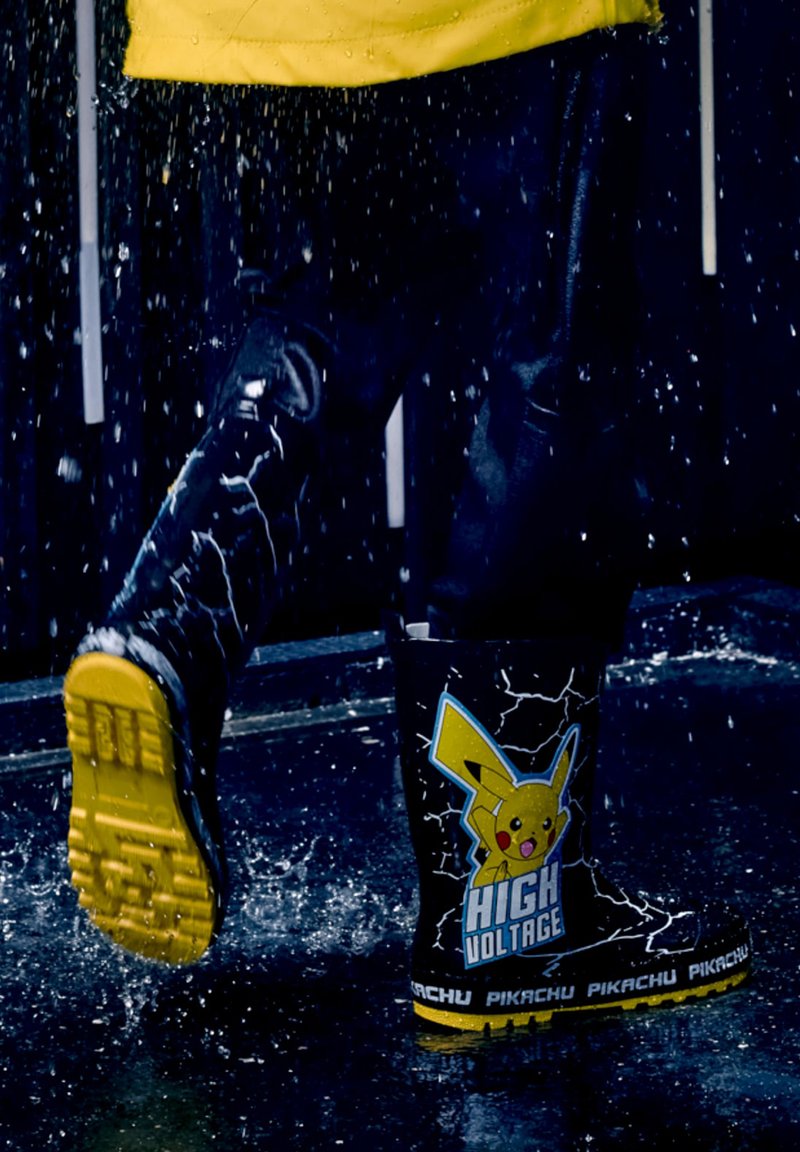 Enfant portant des bottes de pluie noires avec des semelles jaunes et un motif Pikachu «High Voltage» éclaboussant dans une flaque par une journée pluvieuse.
