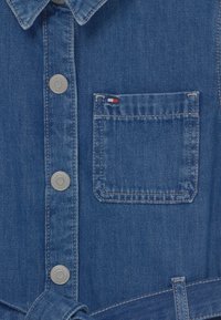 Camisa de mezclilla azul con botones plateados, un bolsillo en el pecho que presenta un pequeño logo rojo, blanco y azul, y un cinturón atado en la parte inferior.