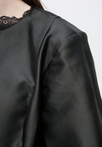 Top din satin negru cu un decolteu delicat din dantelă, textură netedă și cusătură structurată la nivelul umerilor pentru un finisaj elegant.