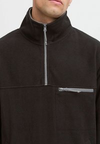 Black fleece pullover with a high collar, featuring a central zip and a small zippered pocket on the chest with gray accents.